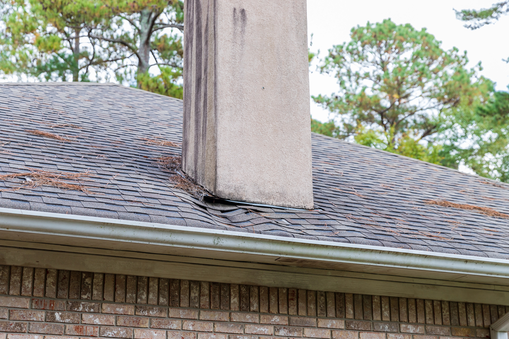 A brick chimney with visible water damage and sagging shingles on a pitched roof with scattered pine needles.