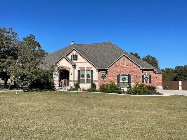 Single-story brick house with a dark gray roof, large front yard, driveway, and several windows decorated with wreaths under a clear blue sky.