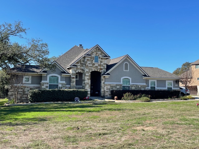 Single-story house with stone and stucco exterior, arched entry, multiple gables, and a large front lawn under a clear blue sky.