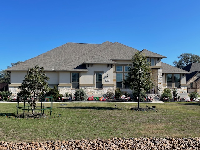 Single-story suburban house with a gray roof, stone and stucco exterior, landscaped front yard, small trees, and decorative plants, under a clear blue sky.