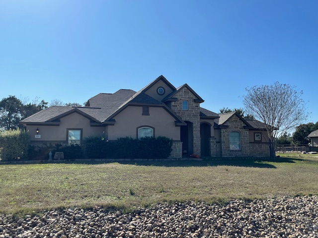 Single-story suburban house with tan stucco and stone exterior, dark roof, bushes in front, a leafless tree on the right, and a rocky foreground under a clear blue sky.