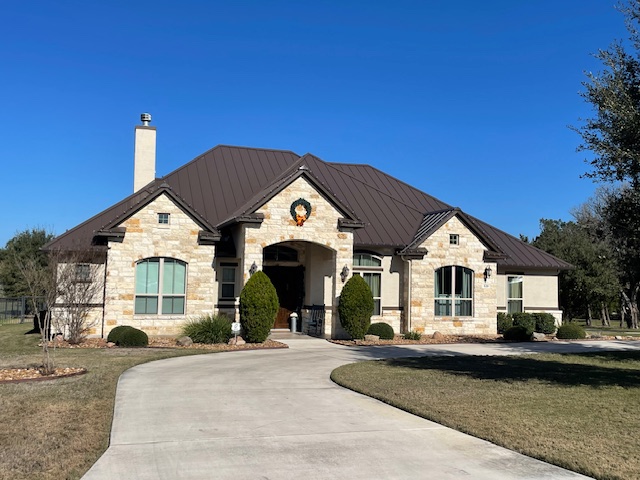 Single-story stone house with a dark metal roof, arched windows, a wreath above the entrance, manicured bushes, and a curved driveway under a clear blue sky.